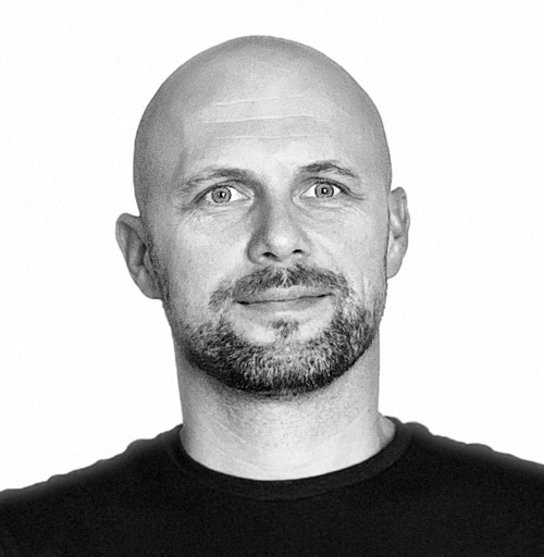 A bald man with a beard and mustache is looking straight ahead. He is wearing a dark shirt, and the background is white, capturing a moment that feels suspended in time.