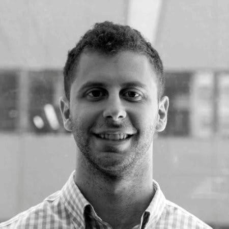 Black and white photo of a person with short curly hair wearing a checkered shirt, smiling at the camera with a background of large windows, capturing a genuine moment in the real world.