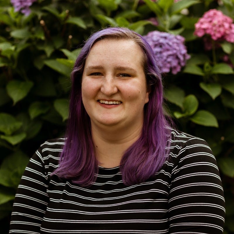 A woman with purple hair standing in front of flowers, showcasing the observability trend in 2024.