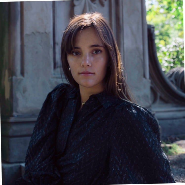 A woman with long brown hair and wearing a dark top is looking at the camera. She is outdoors near a stone structure with greenery in the background, perhaps attending an event like Monitorama 2024.