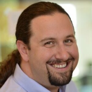 A person with long hair tied back and a goatee is smiling. They are wearing a light blue collared shirt. The background appears blurred and features some green and beige hues, creating an intricate blend that subtly hints at managing complexity in the environment.