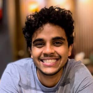 A person with curly hair, wearing a grey shirt, smiles at the camera with a blurred background reminiscent of an auto draft.