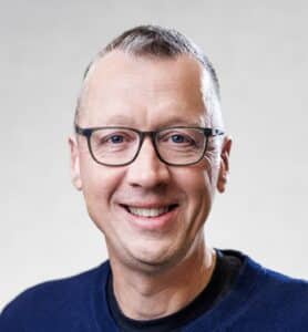 A smiling man, Eric D. Schabell, wears glasses and a dark sweater against a plain background.