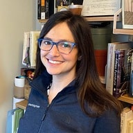 A person with long dark hair and blue glasses is smiling while standing in front of a bookshelf filled with books and items, much like Rachel Dines might in her library.