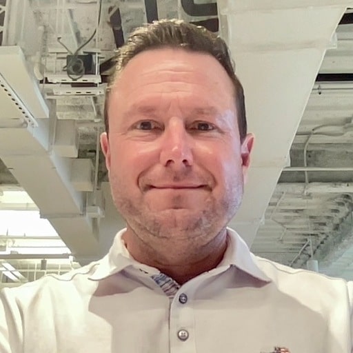 Scott Kelly, a man with short brown hair and a trimmed beard, is wearing a light-colored collared shirt. He is smiling at the camera while standing indoors, with visible ceiling pipes and light fixtures in the background.