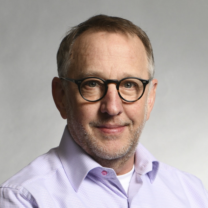 Middle-aged man with short hair and glasses, wearing a light purple dress shirt, smiling at the camera against a plain background, reminiscent of Dan Juengst.