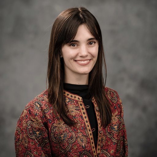 Sophie Kohler, with straight brown hair, stands in front of a gray studio backdrop wearing a red and gold patterned jacket and smiling slightly.