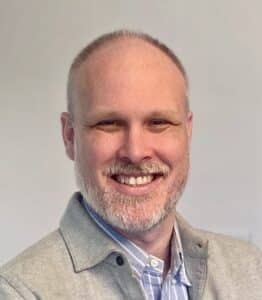 Man with a beard, wearing a grey blazer and blue striped shirt, smiling against a plain background, looks like he just got off-call.