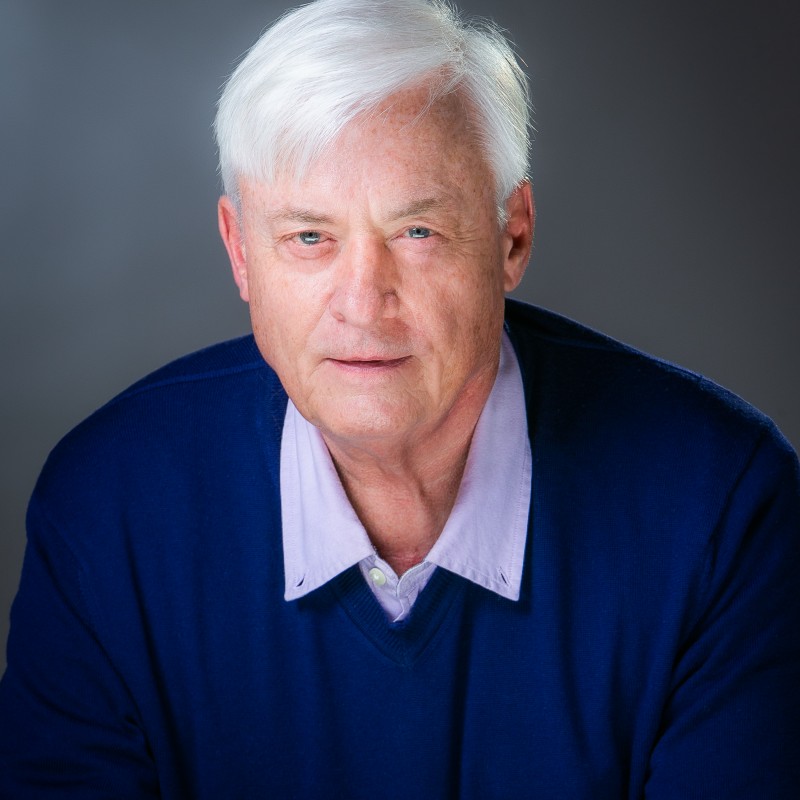 An older man, Eric Newcomer, with white hair and a blue sweater over a light purple shirt stands against a gray background.