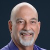 Jason Bloomberg, a person with a bald head and a gray beard, smiles warmly at the camera. He sports a blue shirt that contrasts nicely against the dark background.