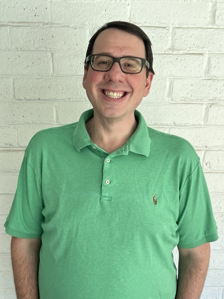 Jacob Rosenberg, wearing glasses and a green polo shirt, smiles warmly while standing against a white brick wall.