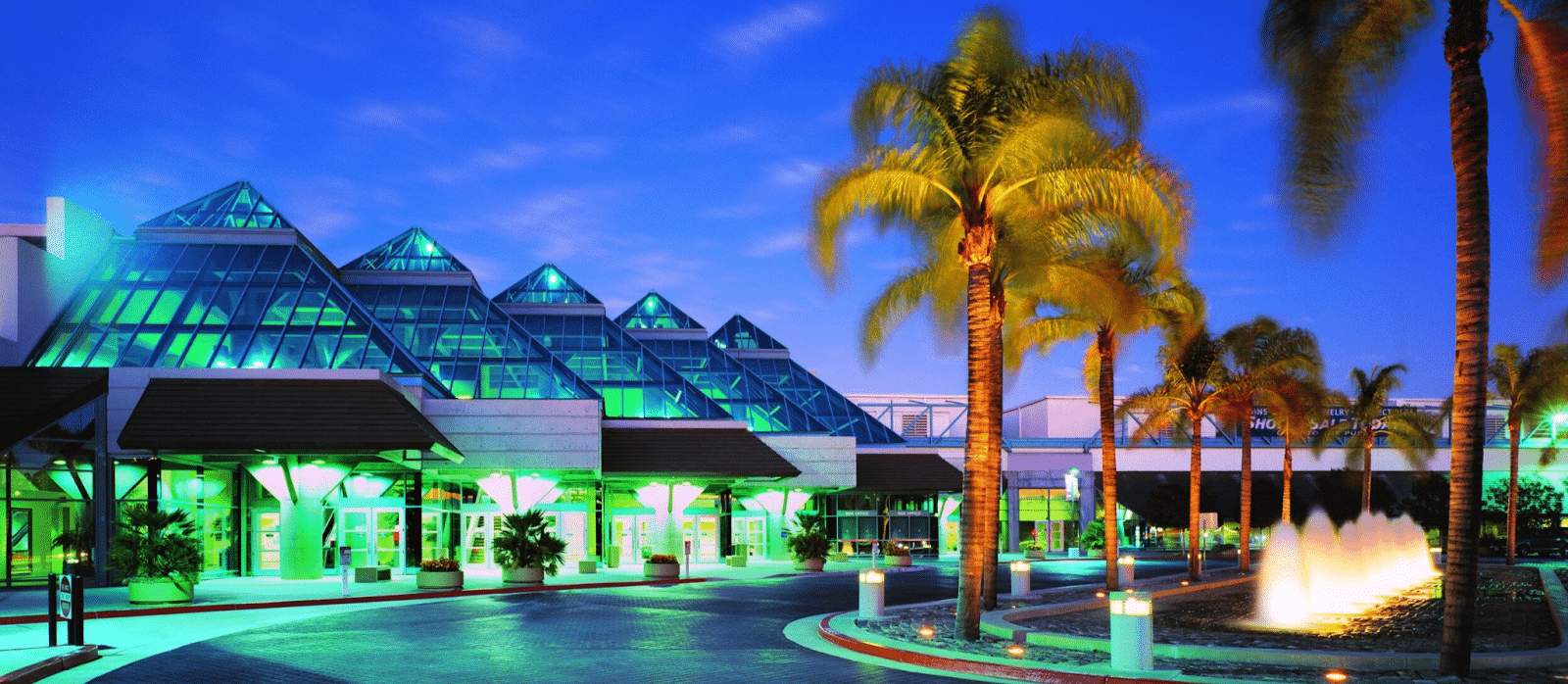 Exterior view of a modern building with glass pyramid structures, surrounded by palm trees and illuminated by green lights, with a lit fountain in the foreground. Join us at DeveloperWeek 2025 to explore this innovative architectural design.