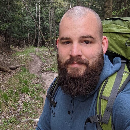 A bearded man in a blue shirt and backpack, resembling a copy of an outdoor enthusiast, stands confidently on the forest trail. Trees and lush greenery form the backdrop to this scene that feels like encountering a new person embracing nature