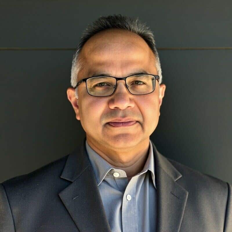 A man with glasses and short hair is wearing a dark blazer and light shirt, standing against a dark background, looking at the camera.