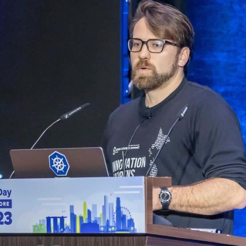 Bryan Oliver, wearing glasses, speaks at a podium with a laptop adorned with a Kubernetes sticker during a tech conference.