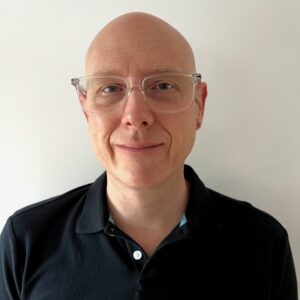 A new person with glasses and a bald head, wearing a black collared shirt, stands confidently against a plain white background.