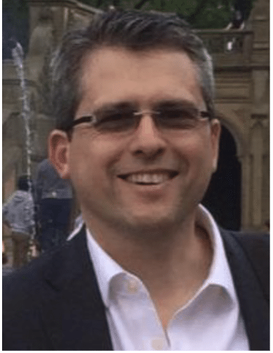 Nic Cheneweth, a smiling man with short dark hair and glasses, dons a dark suit with a white shirt as he stands outdoors before a fountain and stone building.