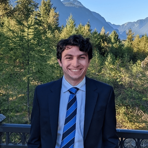 A man in a dark suit, white shirt, and striped blue tie stands outdoors on a balcony with a forest and mountains in the background.