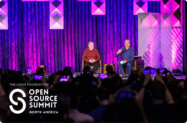 Two speakers sit on stage in front of a seated audience at the Linux Foundation Open Source Summit North America, with purple lighting and geometric stage decor, capturing the collaborative spirit seen at events like the Open Observability Summit.