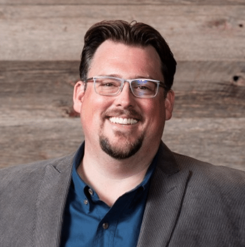 A man with glasses, a beard, and short dark hair is smiling, wearing a blue shirt and a grey blazer, standing in front of a wooden wall.