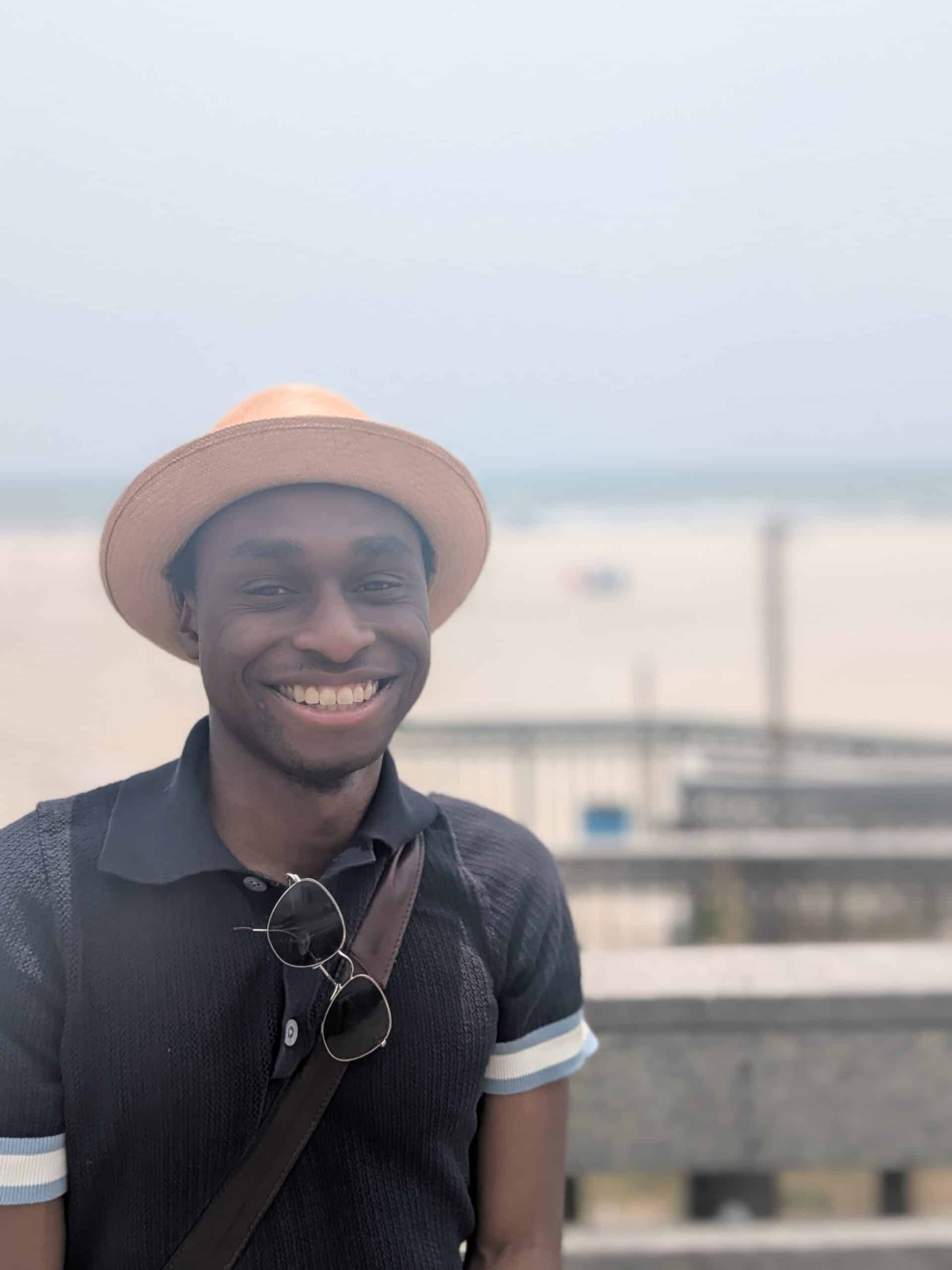 A man wearing a straw hat, sunglasses, and a navy shirt smiles while standing on a boardwalk with a sandy beach in the background.
