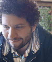 A man with curly hair and a beard, wearing a striped shirt and dark jacket, is seated indoors and looking downward with a slight smile.
