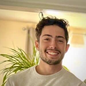 Aissa Mamdouh, a young man with short, curly hair and a beard, smiles while standing indoors near a green plant.