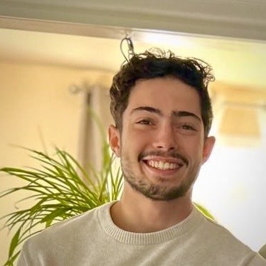 Aissa Mamdouh, a young man with short, curly hair and a beard, smiles while standing indoors near a green plant.
