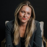 A new person with long blonde hair, wearing a dark blazer, sits in front of a dark background, looking at the camera with a slight smile.