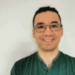 Man with glasses and dark hair tied back, wearing a green shirt, smiling in front of a plain white background.