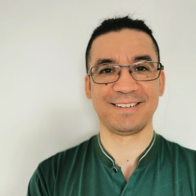 Man with glasses and dark hair tied back, wearing a green shirt, smiling in front of a plain white background.