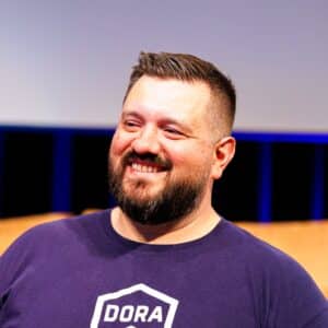A bearded man with short hair smiles while wearing a navy blue "DORA" t-shirt, standing indoors with a blurred background