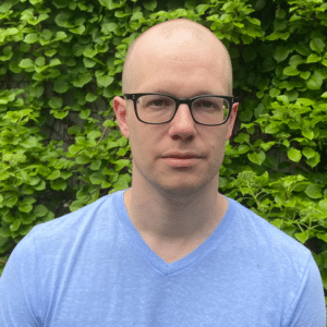 A person with glasses and a shaved head wearing a light blue shirt stands in front of a leafy green background, embodying the thoughtful expertise one might expect from someone helping a company build its own AI SRE.