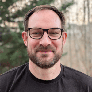 A man with short brown hair, beard, and glasses smiles at the camera while standing outdoors with trees in the background, perhaps envisioning how to build its own AI SRE for a company.
