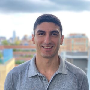 A man with short dark hair, wearing a gray collared shirt, stands outside with blurred buildings and a cloudy sky in the background, perhaps preparing to discuss AI SRE topics for his company’s podcast.
